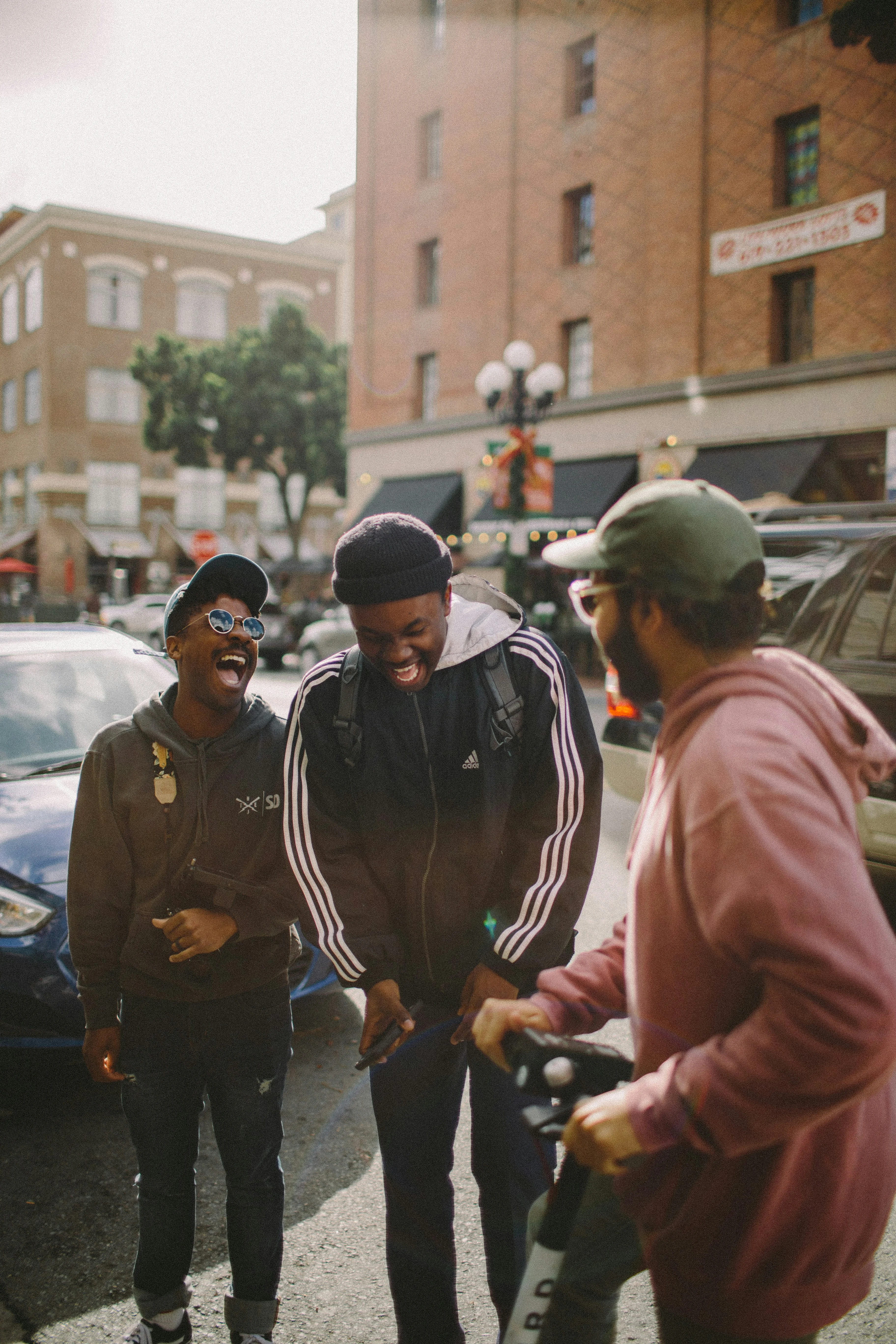 Three men laughing together on a sunlit city street, one doubled over mid-laugh