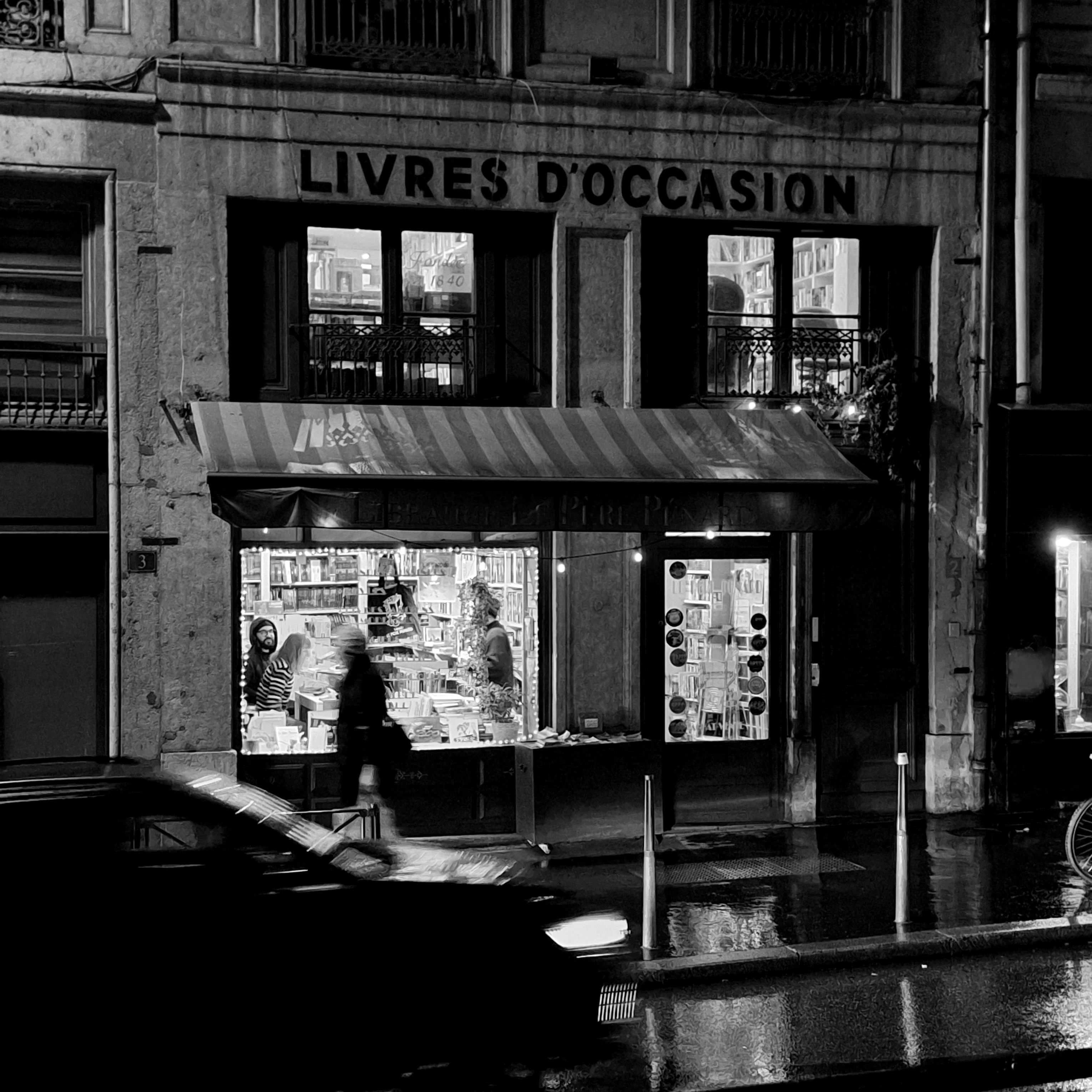 A secondhand bookshop storefront at night, warm light spilling through the window onto a wet street, a person walking past blurred by motion