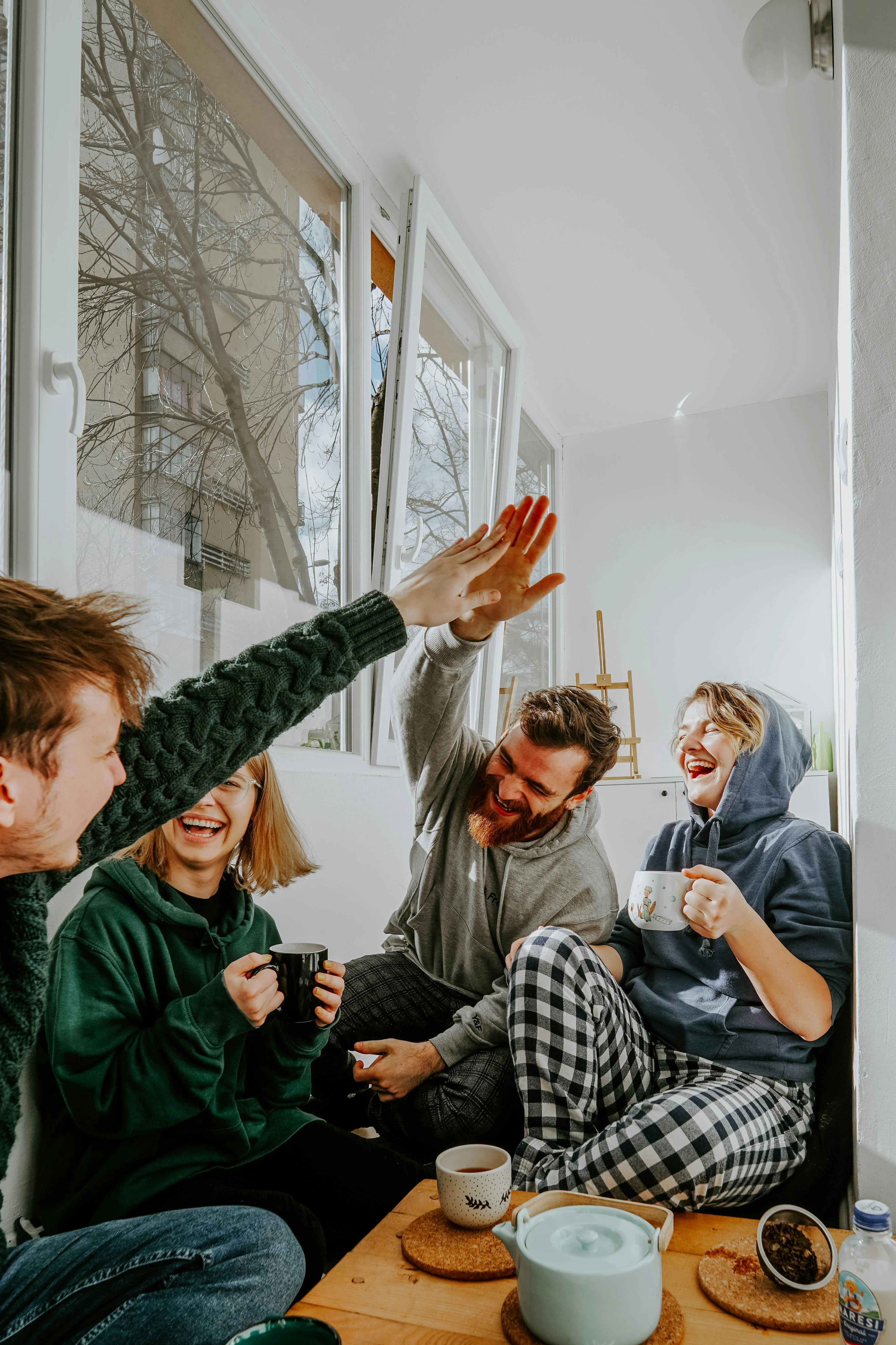A mixed group of friends laughing together over coffee in a bright room, one raising a hand mid-gesture